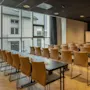 Foredragsstoler vender mot en projektorskjerm i et moderne konferanserom med beige vegger og lyse tregulv. Store vinduer viser en bygning utenfor. Bordene har notatblokker og penner.
