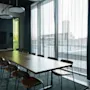 A large wooden conference table surrounded by chairs sits under modern ceiling lights in a room with floor-to-ceiling windows and sheer curtains, creating a bright and spacious setting.