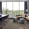 Et konferanserom har et bord med svarte stoler og en vase med blomster. Ved siden av er en sittegruppe med sofaer, puter og et vindu med utsikt over trær.