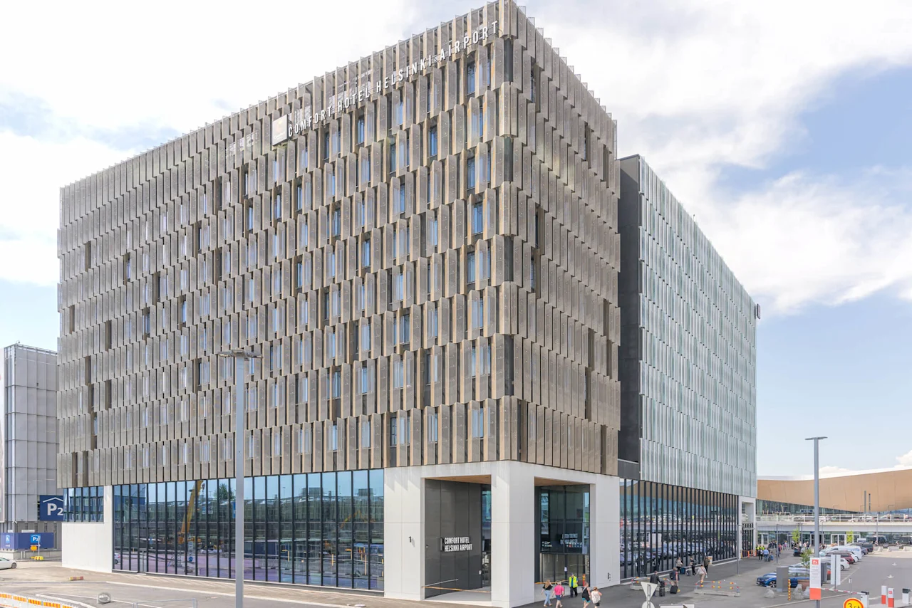 En moderne, geometrisk bygning merket "COMFORT HOTEL HELSINKI AIRPORT" står ved et parkeringsområde under en delvis skyet himmel. Folk går i nærheten.