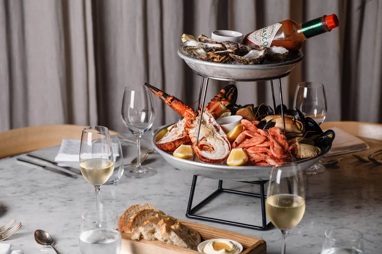 Et etasjefat med sjømat har hummer, reker, østers og sitronbåter på et marmorbord med glass hvitvin og brød. En flaske merket "Tabasco Original Red Sauce" hviler på toppen.