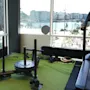 A weighted sled sits on artificial grass indoors, with large windows showing a cityscape and water outside. A wall clock reads 1:15.