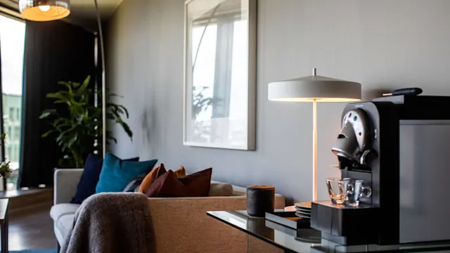Kaffemaskin plassert på et glassbord, ved siden av en moderne lampe, med en sofa dekorert med fargerike puter i en koselig, velinnredet stue med store planter.