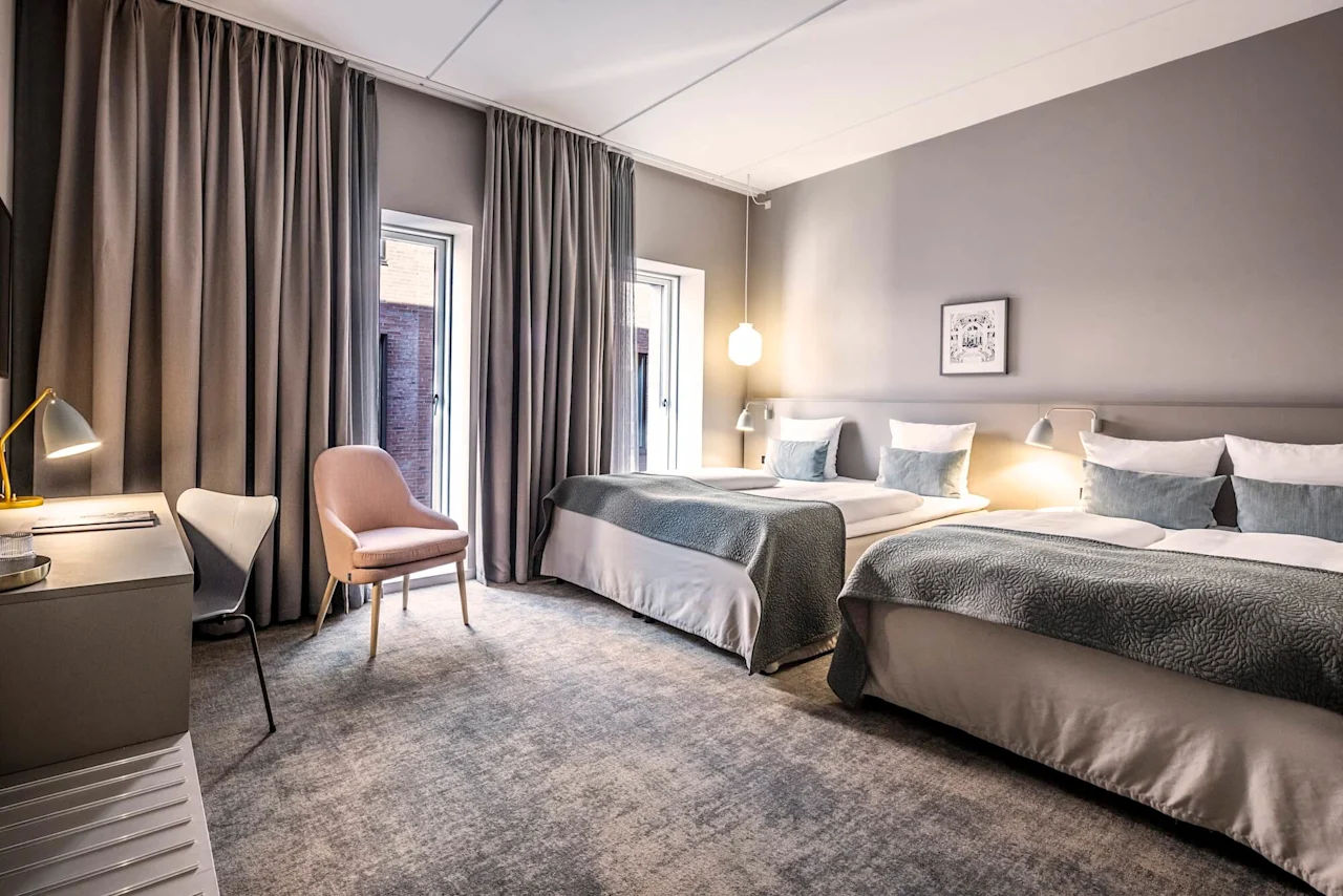 Två prydligt bäddade sängar står i ett modernt hotellrum, omgivna av mjuk belysning, ett skrivbord, stolar och golv-till-tak-gardiner, vilket skapar en mysig och inbjudande atmosfär.