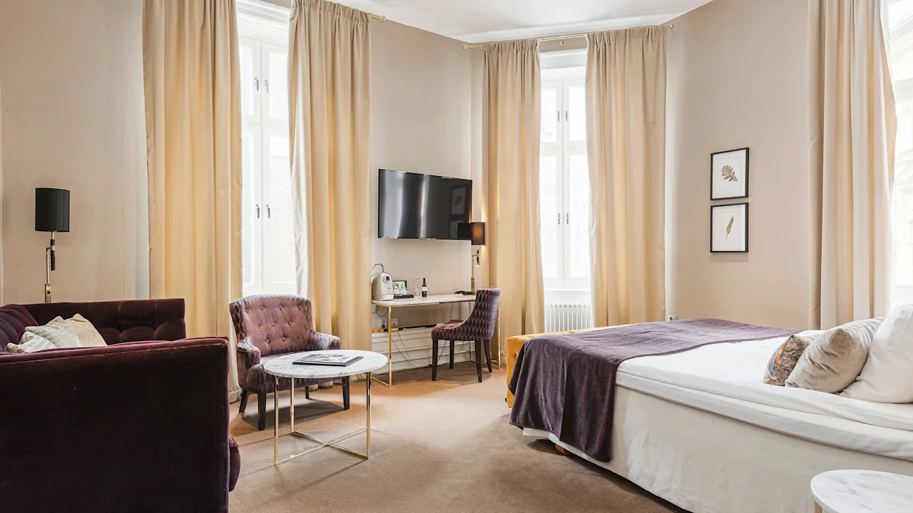 Elegant hotellihuone, jossa on muhkea sohva, nojatuoli, työpöytä ja sänky neutraaleissa sävyissä.