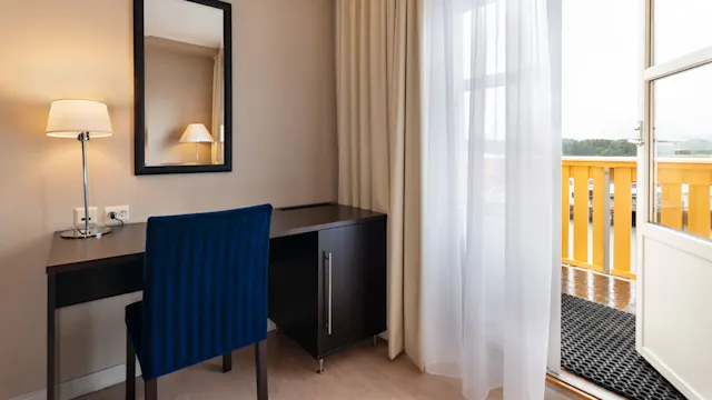 Et sort skrivebord med en blå stol står i et rum ved siden af en lampe og a spejl, ved siden af lette gardiner nær en åben dør ud til en balkon med gult rækværk.