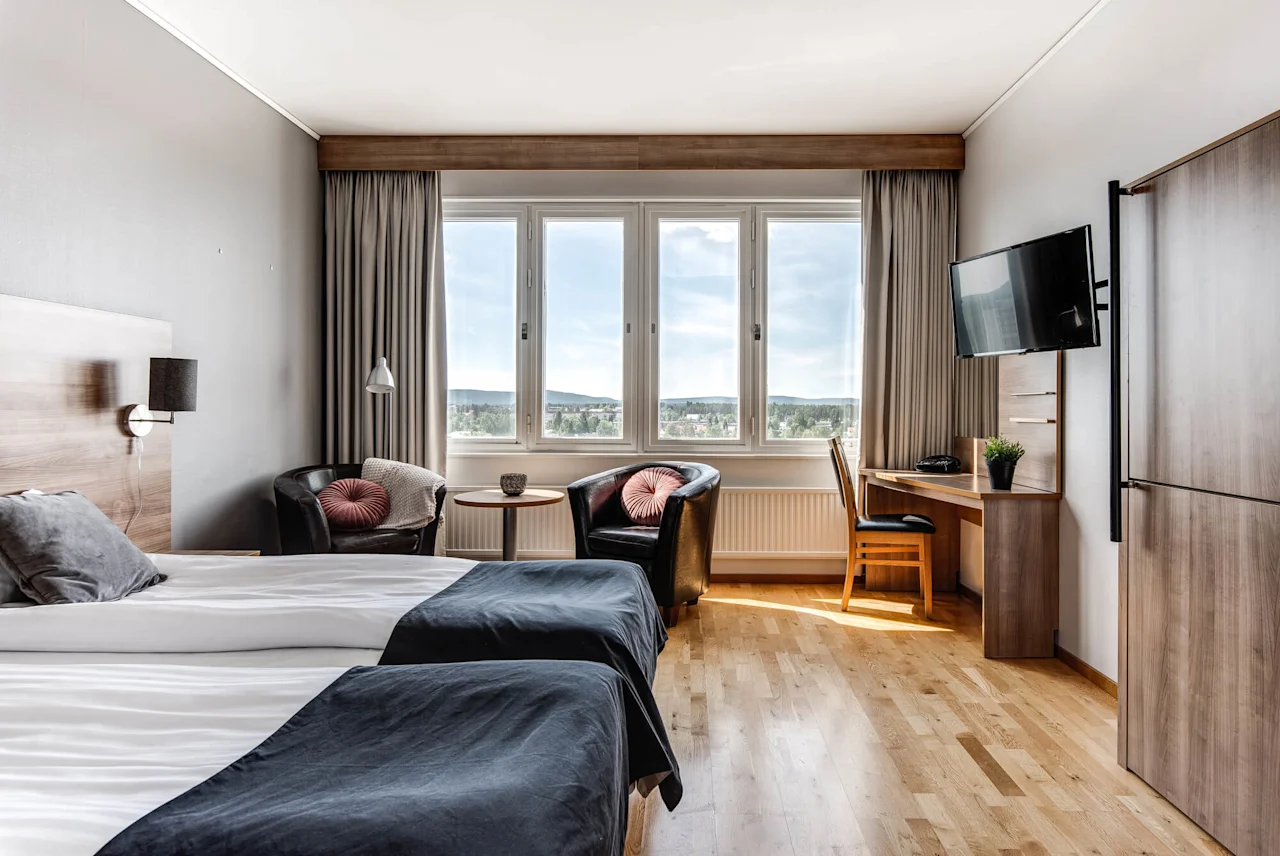 Ett prydligt hotellrum med två sängar med grå sängkläder, två fåtöljer vid ett fönster, en väggmonterad TV och ett skrivbord med en stol med utsikt över en naturskön vy.