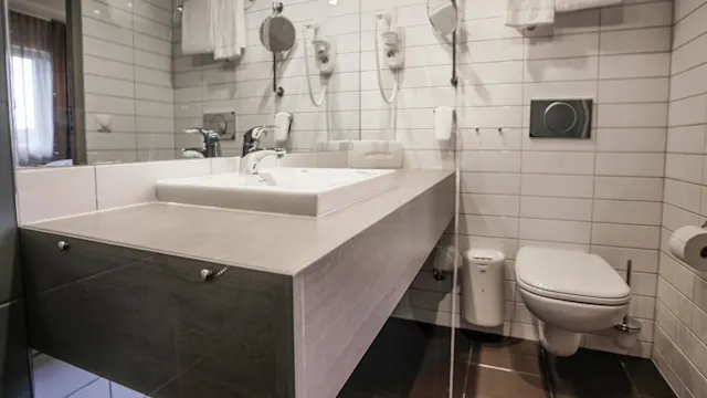 Et moderne bad har en hvit vask og kran på en trebenk, med veggmontert toalett, håndklær og speil mot flislagte vegger under mykt lys.