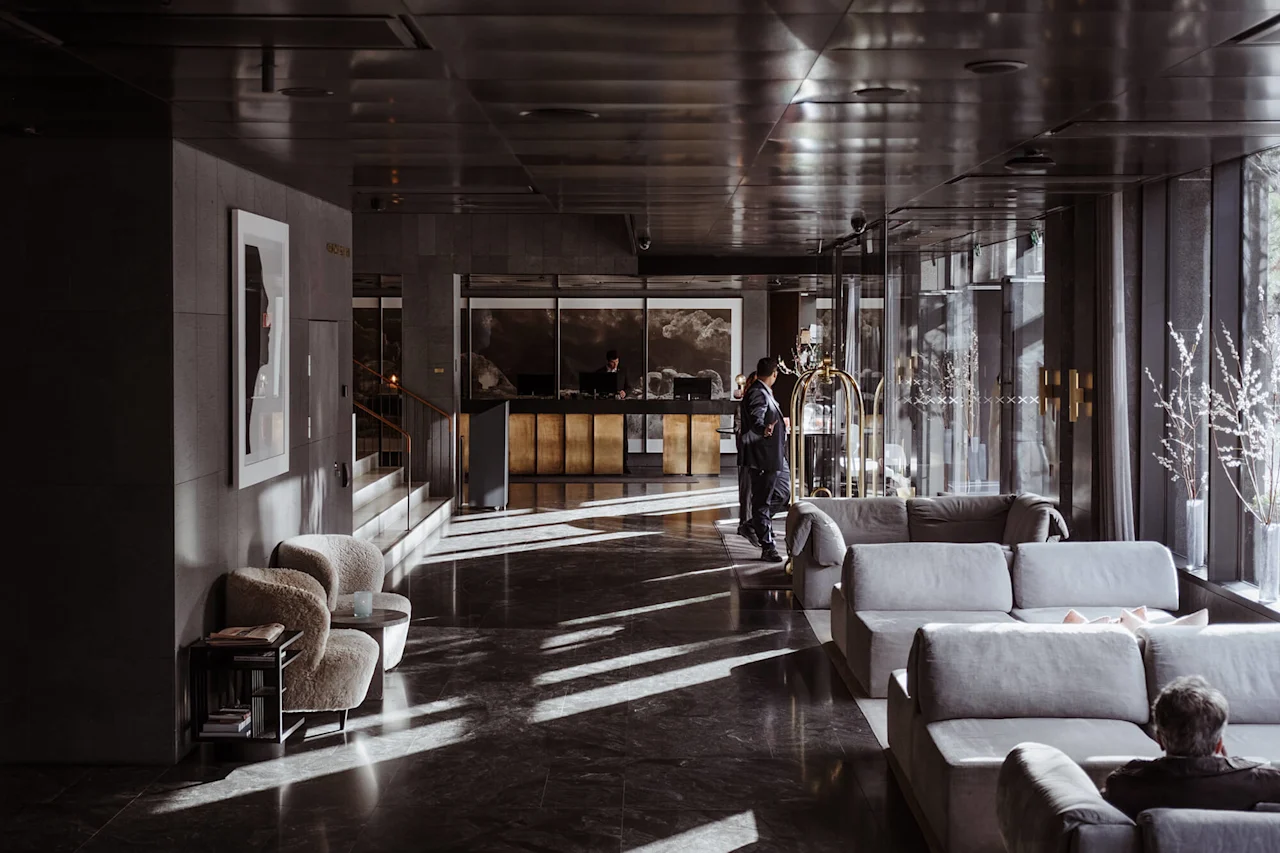 En hotellobby med en resepsjonsskranke; en person sjekker inn. Myke sofaer og lenestoler fyller det solbelyste rommet, med moderne dekor og utsikt fra vinduer.