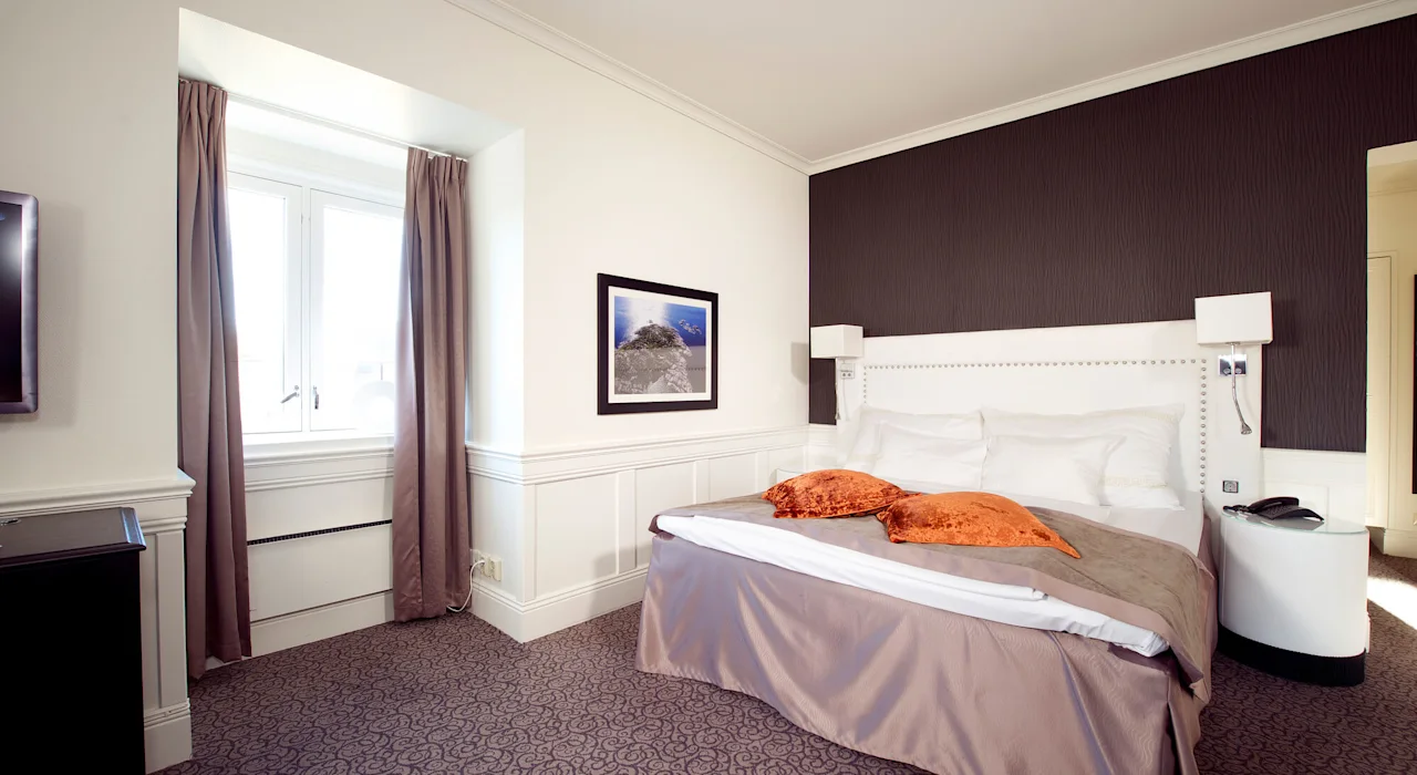 A double bed with white pillows and orange cushions is set against a dark accent wall, flanked by bedside tables, in a hotel room with a window and framed wall art.