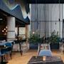 Elegant salong med moderne møbler, sorte bord omgitt av blå stoler, planter som skillevegger. Store vinduer med gardiner, ambient lys, blå bølgedesign på veggen.