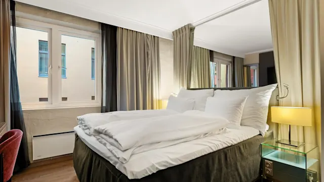 A neatly made bed with white linens is in the center of a cozy room, flanked by curtains, a window, and a bedside table with a lamp.