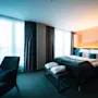 En pent oppredd seng med hvite og blågrønne puter, dekket av et mørkt teppe, står ved siden av store vinduer med gjennomsiktige gardiner, som skaper en lys og innbydende hotellromatmosfære.
