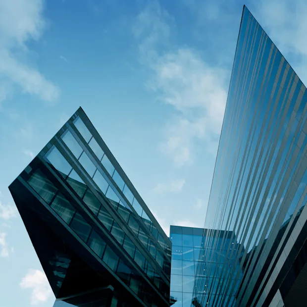 En moderne, vinkel glassbygning strekker seg oppover, og reflekterer den blå himmelen med spredte skyer. Den arkitektoniske designen har skarpe, kryssende linjer og skaper en dynamisk urban estetikk.