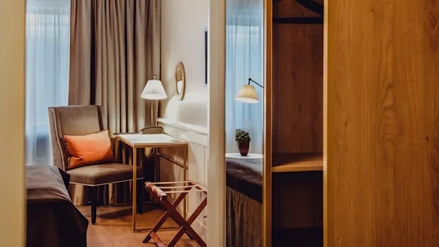 Et koselig rom med en stol, bord, lampe og en sammenleggbar bagasjebrett, varm belysning, og et vindu med lette gardiner.