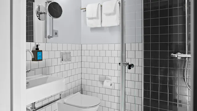 Ett modernt badrum med toalett, handfat och dusch med glasväggar; väggarna är klädda med vita kakelplattor. Handdukar hänger på en ställning. En blå tvålpump är märkt "SUE MUSE".