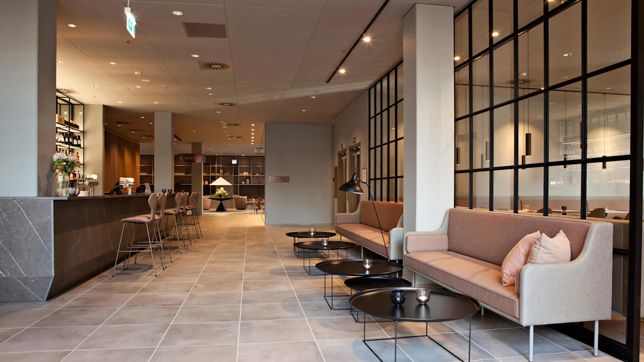 En modern lounge har soffor med kuddar bredvid svarta soffbord; en bar med höga stolar ligger på motsatt sida. Mjuk belysning fyller det eleganta, kaklade rummet.