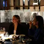 To kvinner sitter ved et svakt opplyst bord, en drikker fra et glass, den andre ser framover. Dekorative glasspaneler og en mykt glødende lampe skaper en koselig restaurantatmosfære.