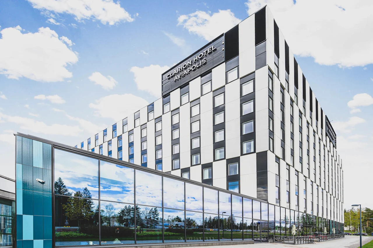 En fleretasjes hotellbygning med rutefatature står under en blå himmel. Store vinduer reflekterer trær. Tekst: "CLARION HOTEL AVIAPOLIS."