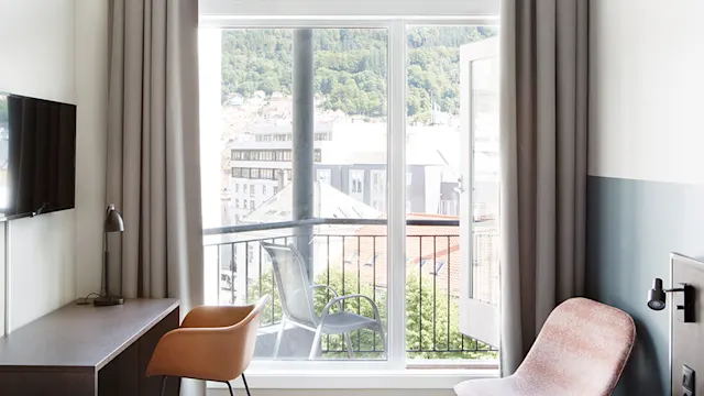 Et lyst hotellrom med skrivebord, lampe, stoler og en veggmontert TV. En åpen dør fører til en balkong med utsikt over hustak og grønne åser.
