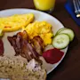 Plate with scrambled eggs, bacon, omelet, sliced cucumber, beet puree, and bread, served with a glass of orange juice on a wooden table.