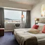 Et hotellrom har en seng med dekorative puter, et skrivebord med en stol og et stort vindu med utsikt over åser og vann.