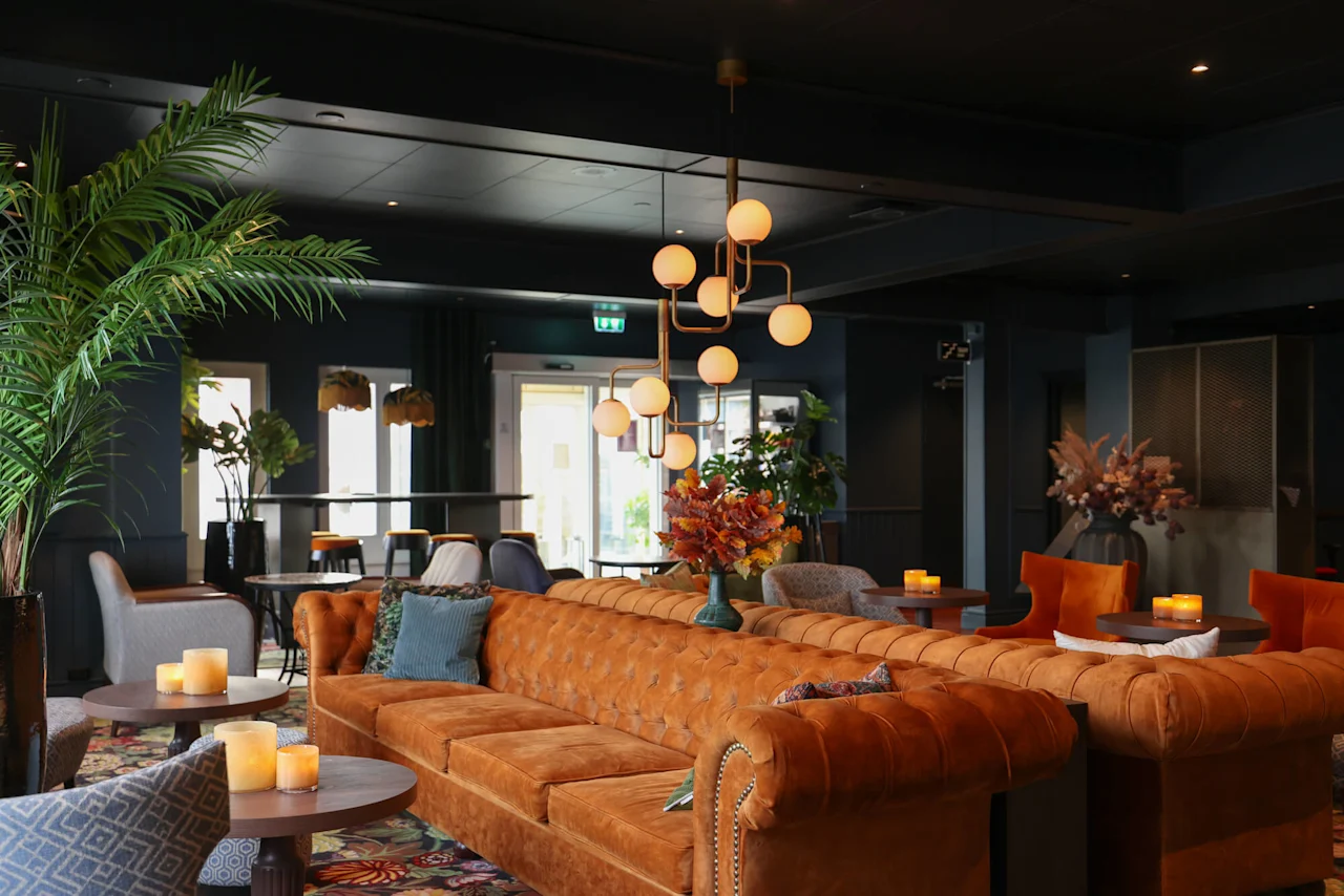 En brun lædersofa med puder står under en moderne lysekrone, omgivet af potteplanter, stole og borde med lys i en elegant indrettet lounge.
