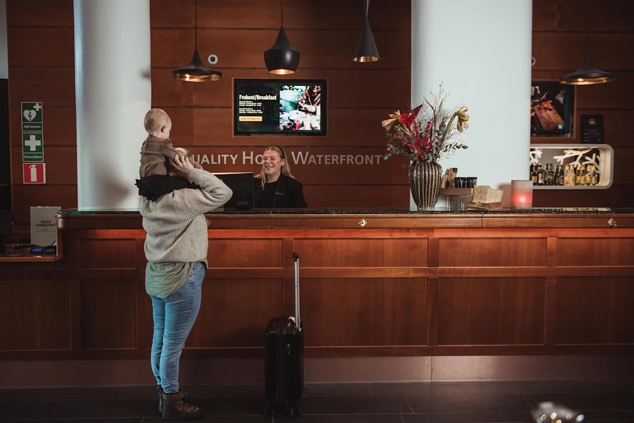 Henkilö kantaa lasta olkapäillään hotellin vastaanotossa. Vastaanottovirkailija hymyilee tiskin takana. Seinällä lukee "QUALITY HOTEL WATERFRONT". Lähellä on matkalaukku ja kukkia. Teksti: "Frukost/Breakfast".