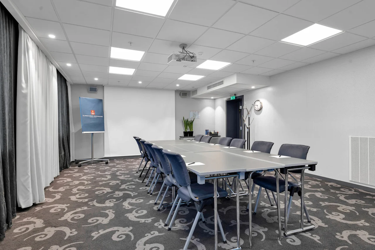 A rectangular conference table is surrounded by blue chairs in a carpeted meeting room. A whiteboard with the text "CLARION HOTEL TREND STANDARD" stands beside curtains.