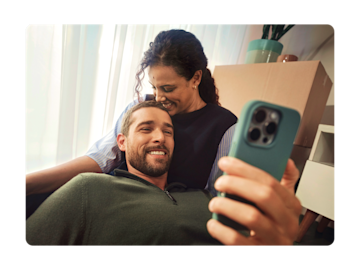 A couple sits smiling together while looking at a smartphone.