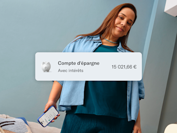 Femme vêtue de bleu, assise et tenant son téléphone portable avec l'application N26 ouverte. Au premier plan, solde du compte principal et du compte d'épargne.