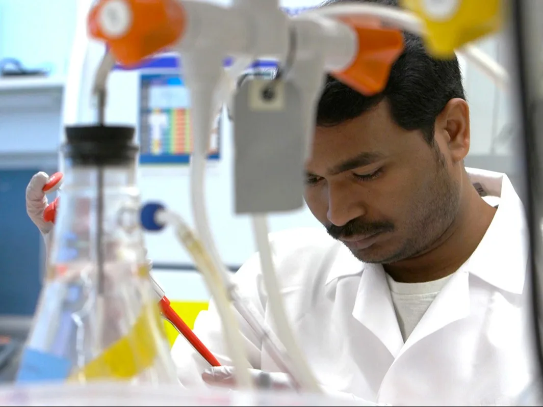 A researcher looks down while working in a lab; laboratory equipment is in the foreground.