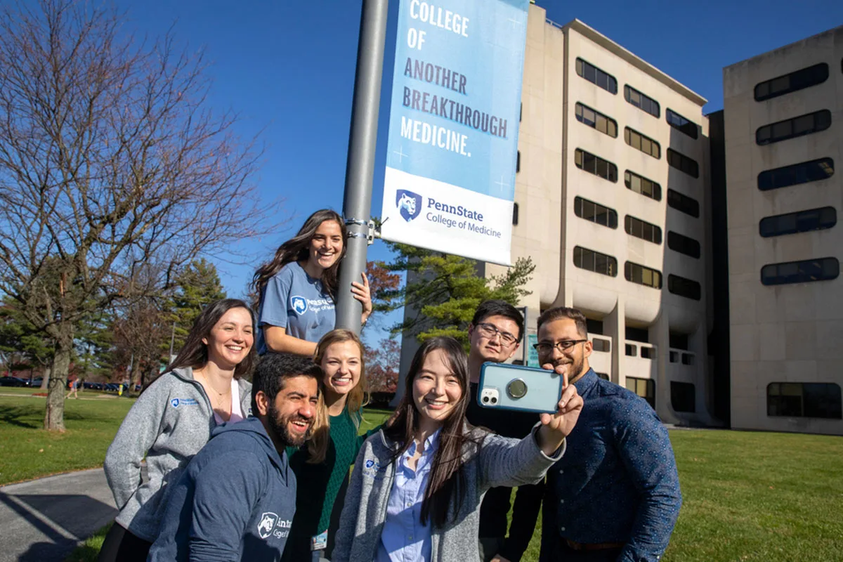Seven students take a selfie by a College of Medicine banner outside the school's crescent building in Hershey
