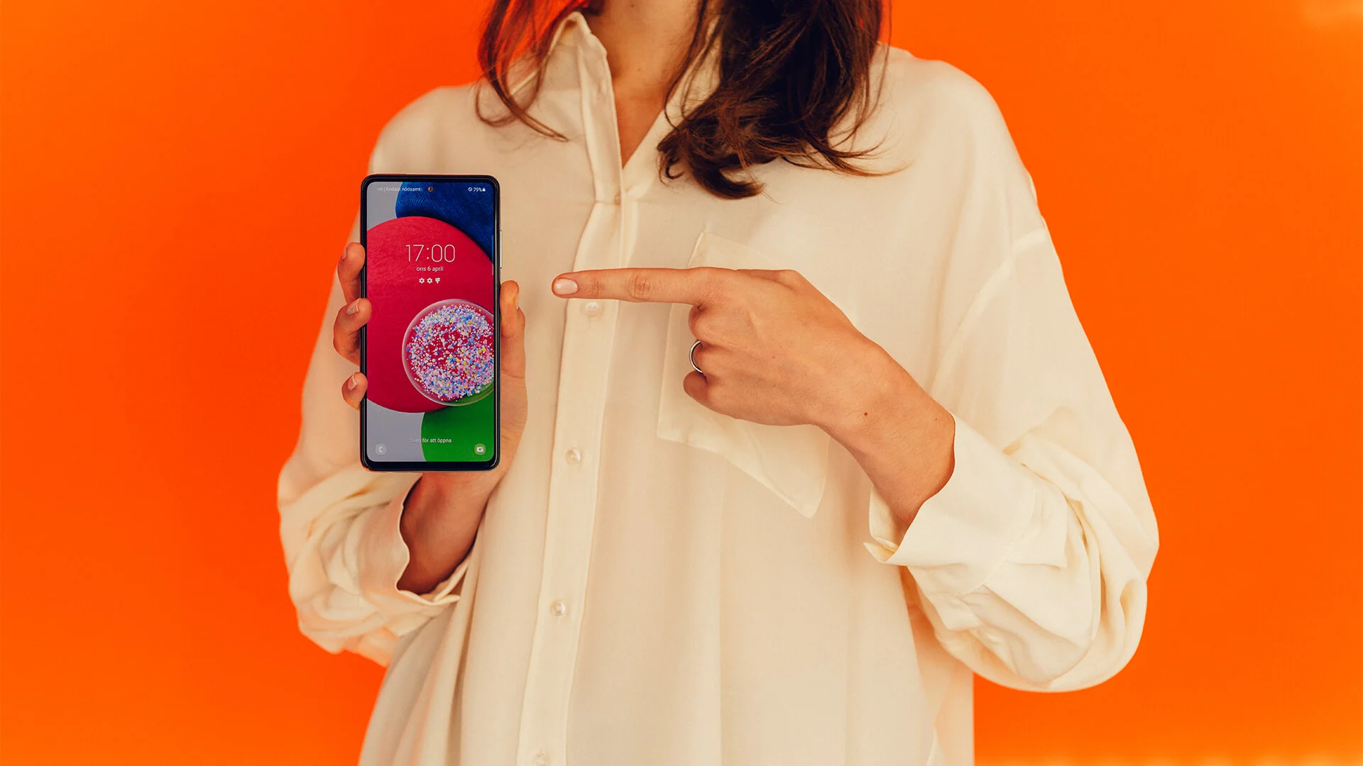 Kvinna i vit skjorta står med en androidmobil i handen och pekar på den med den sin vänstra hand. Bakgrunden är knallorange.