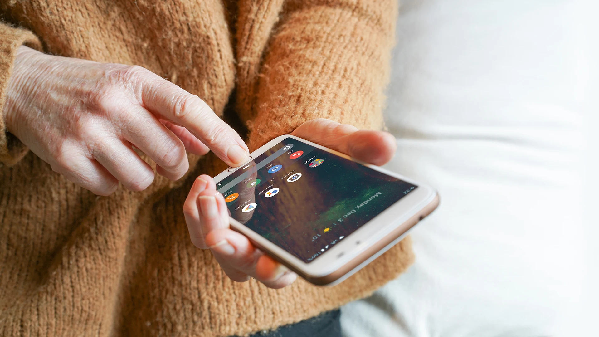 Mobil som är en smartphone hålls i handen, personen har en orange stickad tröja. Personen trycker på huvudknappen med sitt pekfinger. 