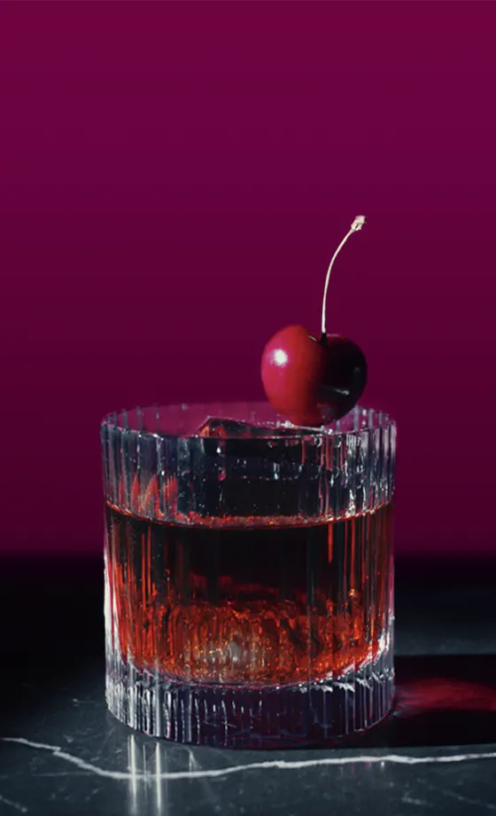 Un vaso con líquido rojo oscuro con un gran cubo de hielo y una cereza en el borde. El vaso tiene un patrón texturizado y está colocado sobre una superficie oscura y un fondo magenta que contrastan.