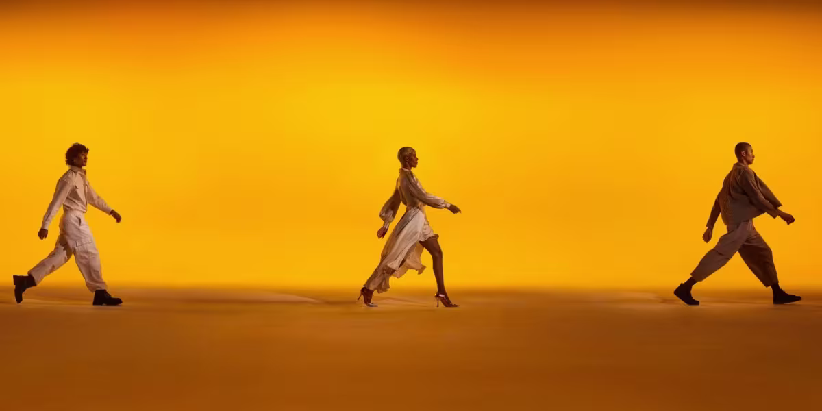 Three people walk in profile against a vibrant orange backdrop. Each is dressed in earth-toned, flowing outfits, creating a sense of motion and style within a minimalist setting.