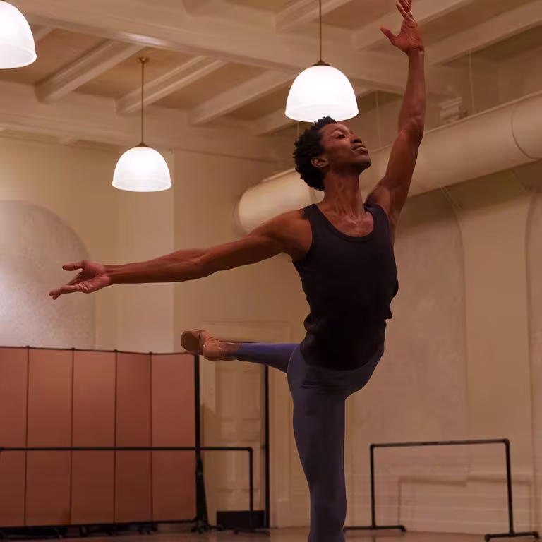 A ballet dancer in a black tank top and blue leggings performs an elegant arabesque in a spacious dance studio with high ceilings and hanging lights. The room features a folding screen and barre in the background.