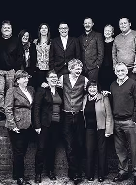 Black and white photo of a group of thirteen people standing and smiling. They are dressed in formal and semi-formal attire, and they are posing against a dark background.
