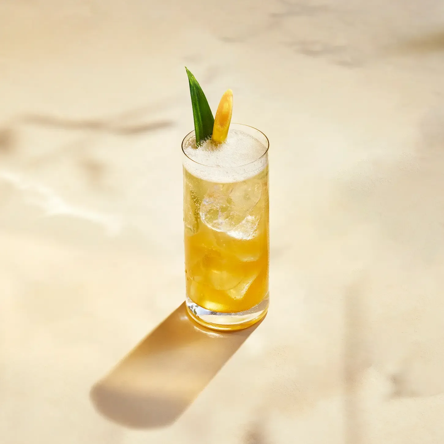 Un vaso alto lleno de un cóctel amarillo, cubitos de hielo, una capa espumosa y adornado con una hoja de piña y una cáscara de limón. Proyecta una sombra sobre una superficie clara.