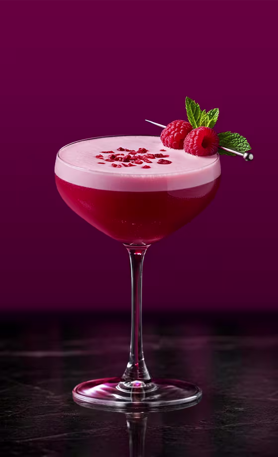 Un cóctel rojo vibrante en copa coupé, coronado con una capa rosa espumosa y decorado con frambuesas y menta. El fondo es de un magenta intenso y profundo.