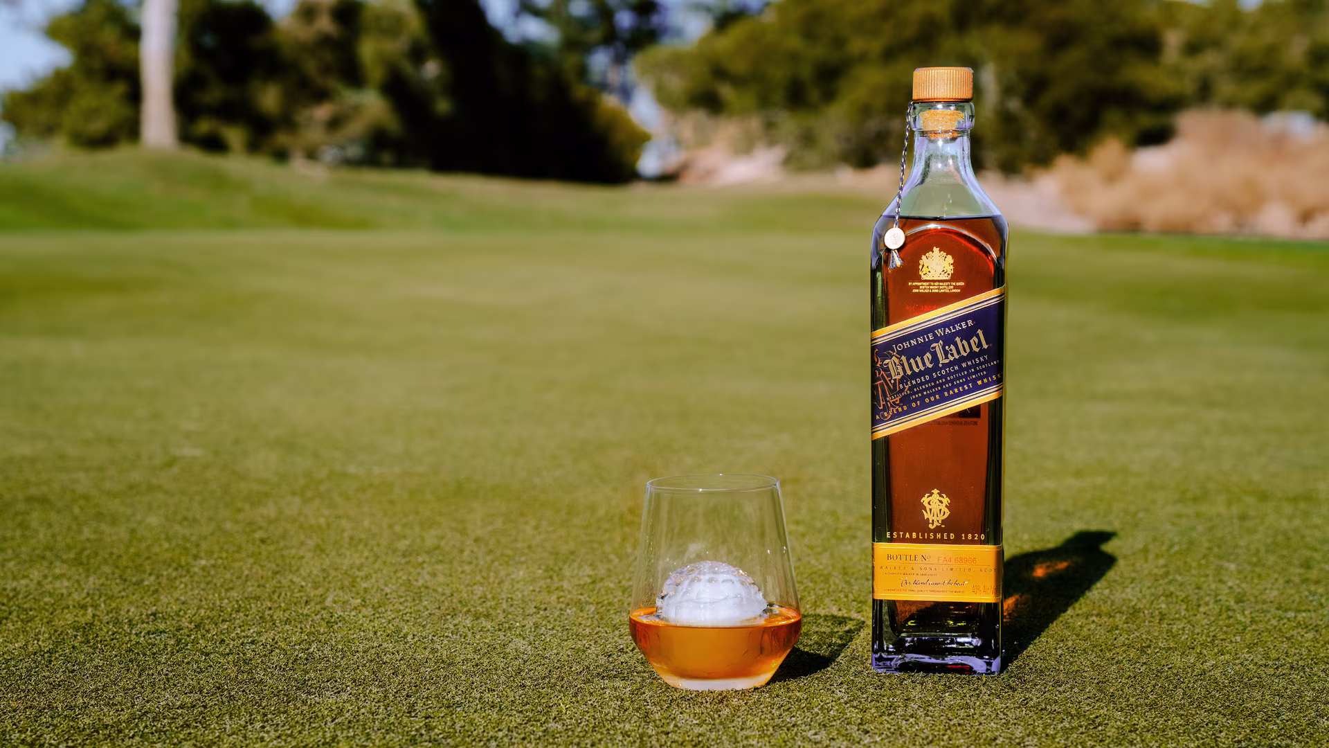 Una botella de whisky Johnnie Walker Blue Label reposa sobre el césped junto a un vaso de whisky con una pelota de golf dentro. Al fondo se ven árboles y un campo de golf borroso.