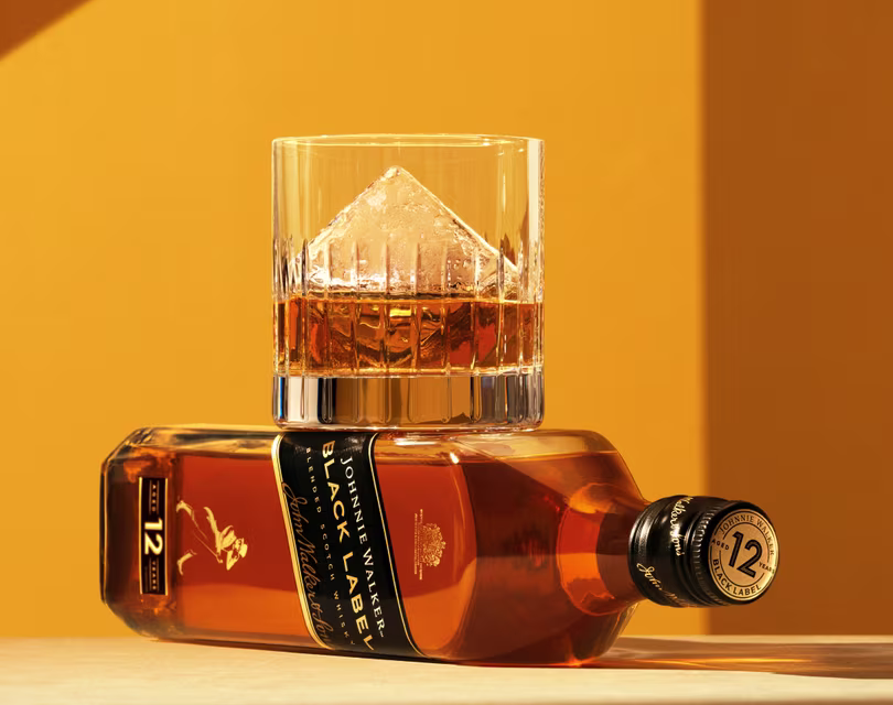 Un vaso de whisky con un gran cubo de hielo con forma de pirámide reposa sobre una botella horizontal de whisky escocés Johnnie Walker Black Label de 12 años sobre un fondo amarillo cálido.