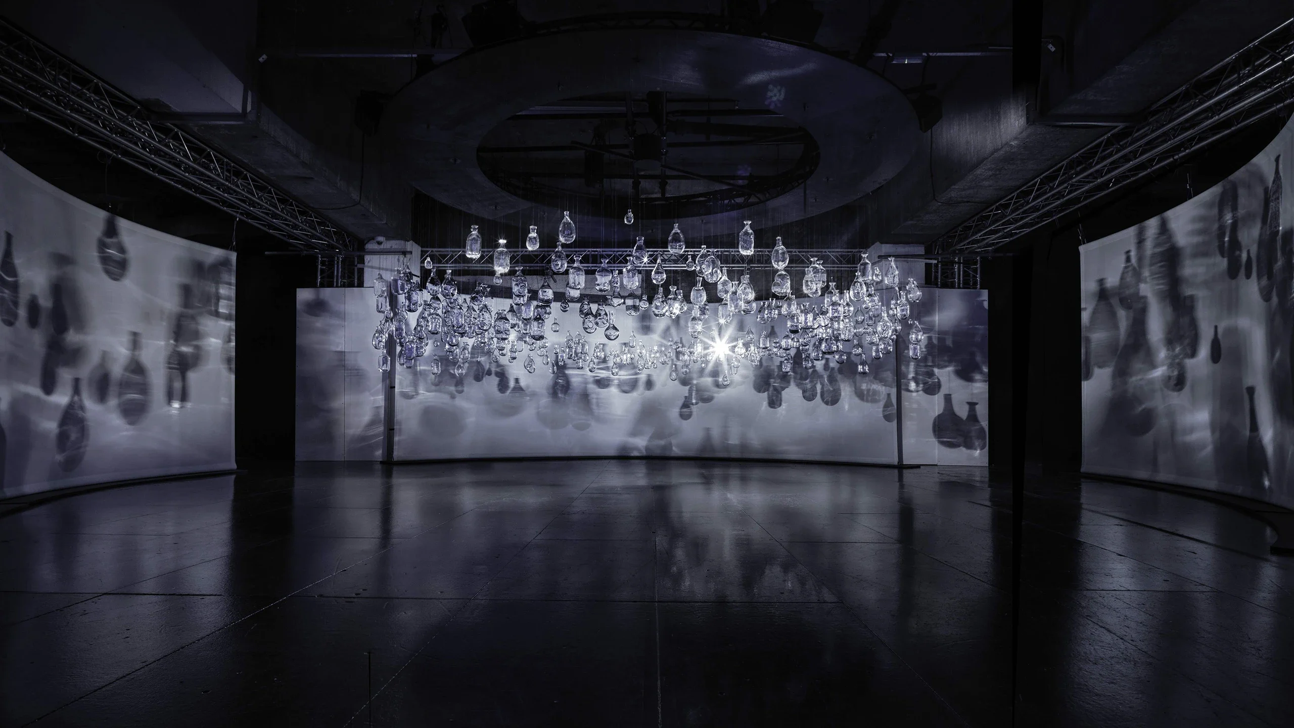 A dark, modern art gallery features numerous transparent glass bulbs suspended from the ceiling, casting intricate shadows and reflections on large curved white screens behind them.