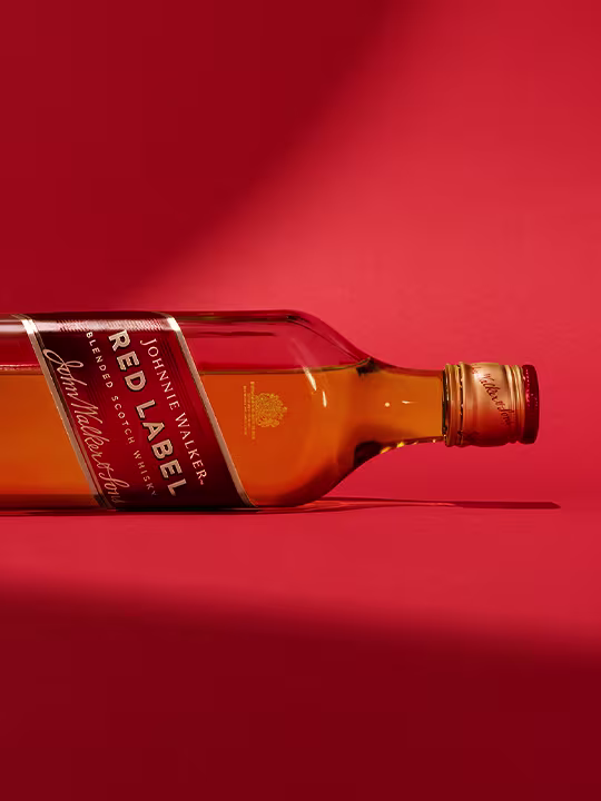 Johnnie Walker Red Label Scotch 위스키 한 병이 빨간색 배경에 옆으로 누워 있습니다. 브랜드의 시그니처 디자인이 특징인 라벨이 보입니다. 투명한 유리잔은 위스키의 호박색을 잘 보여줍니다.