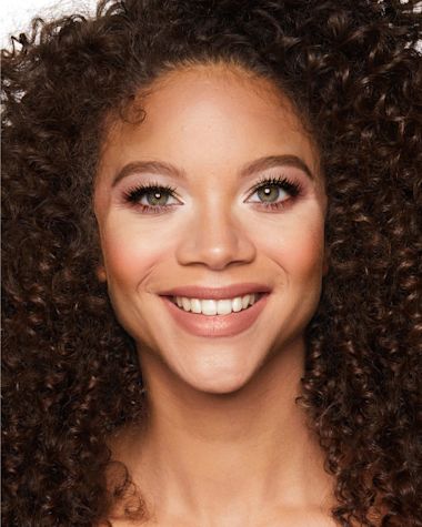 Medium-tone model with green eyes wearing smokey brown and satin-finish peach eyeshadow with glowy peach blush, and subtle champagne highlighter with sandy-taupe bronzed skin. 