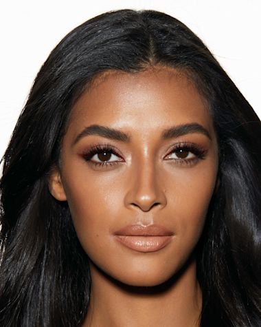 Deep-tone model with brown eyes wearing a nude peach lipstick with a glowy face base, a berry-brown eyeliner in a wing style. 