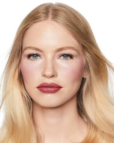 A fair-tone model with blue eyes wearing a lip and cheek cream blush in a glowy deep berry-pink colour on her lips and cheeks. 