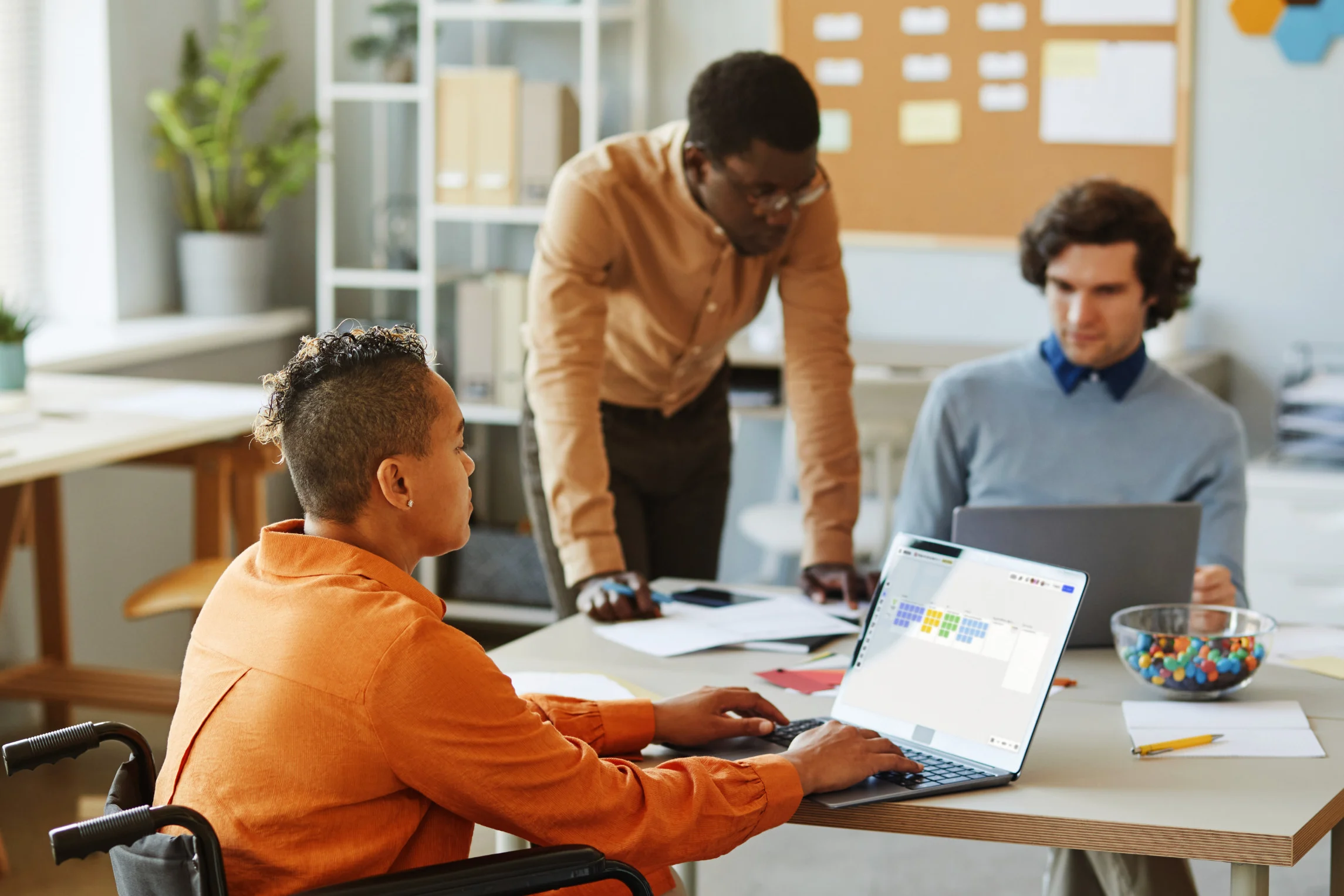 Three colleagues in a casual office setting, collaborating around a table with laptops and documents